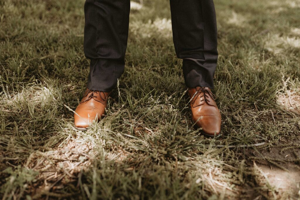 The groom wearing a black suit with brown dress shoes, paired with taupe accents.