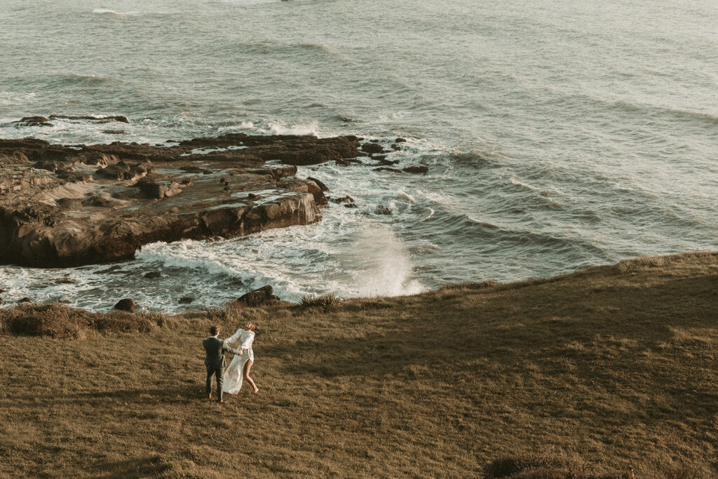 Bride and groom run and play near the coastline during dreamy sunset cliff elopement photos at Crook Point, Oregon.