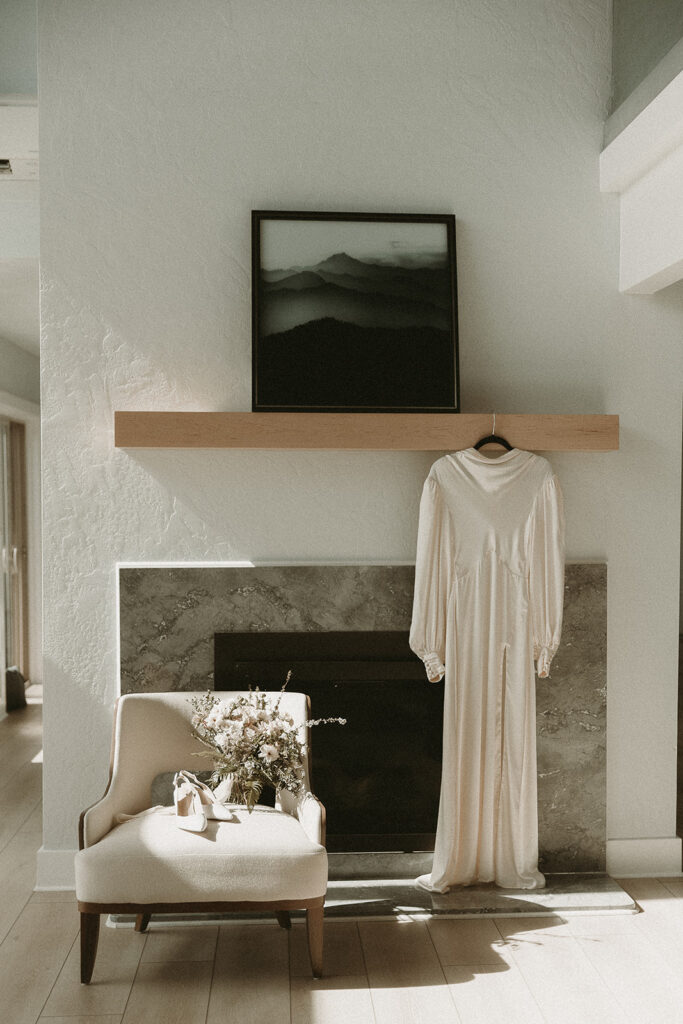 Wedding dress hangs from the mantle beside a chair holding heels and a boquet for an Oregon elopement.
