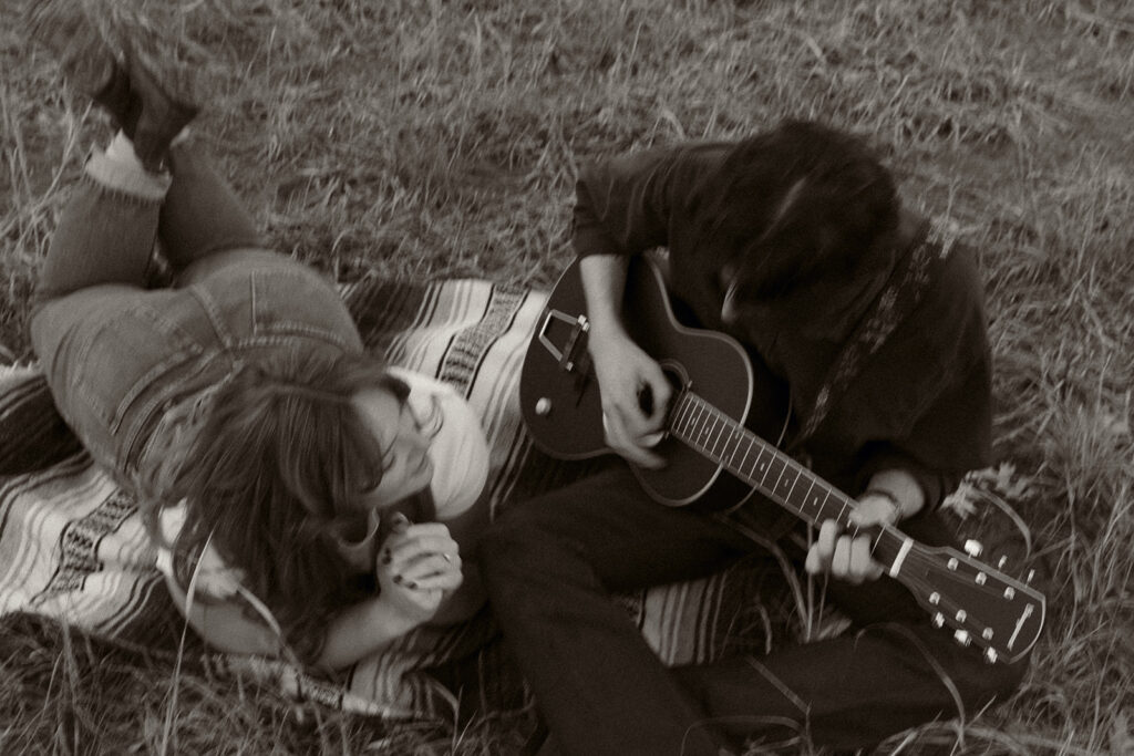 Young man plays guitar for his fiance on a blanket in a bluff field at their engagement session in Kansas City, MO.