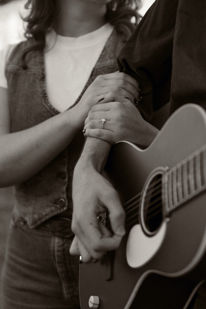 Young woman holds her fiances arm as he plays guitar for her at their engagement session in Kansas City, MO.